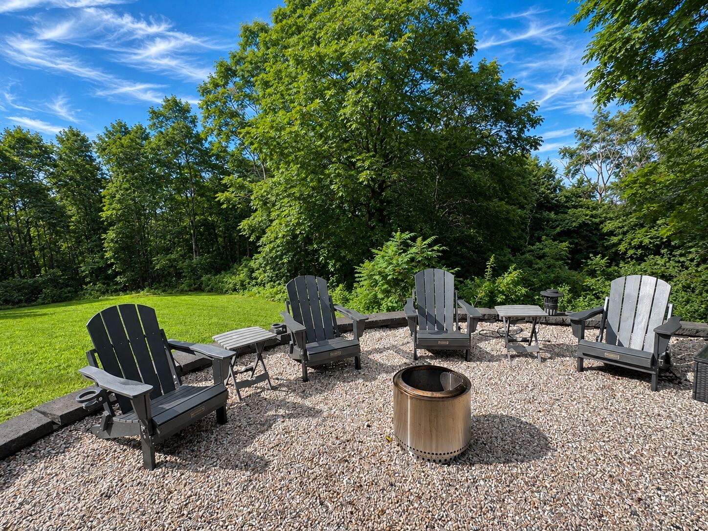 Stone-edged gravel fire pit circle with six Adirondack chairs backing onto lawn and treeline