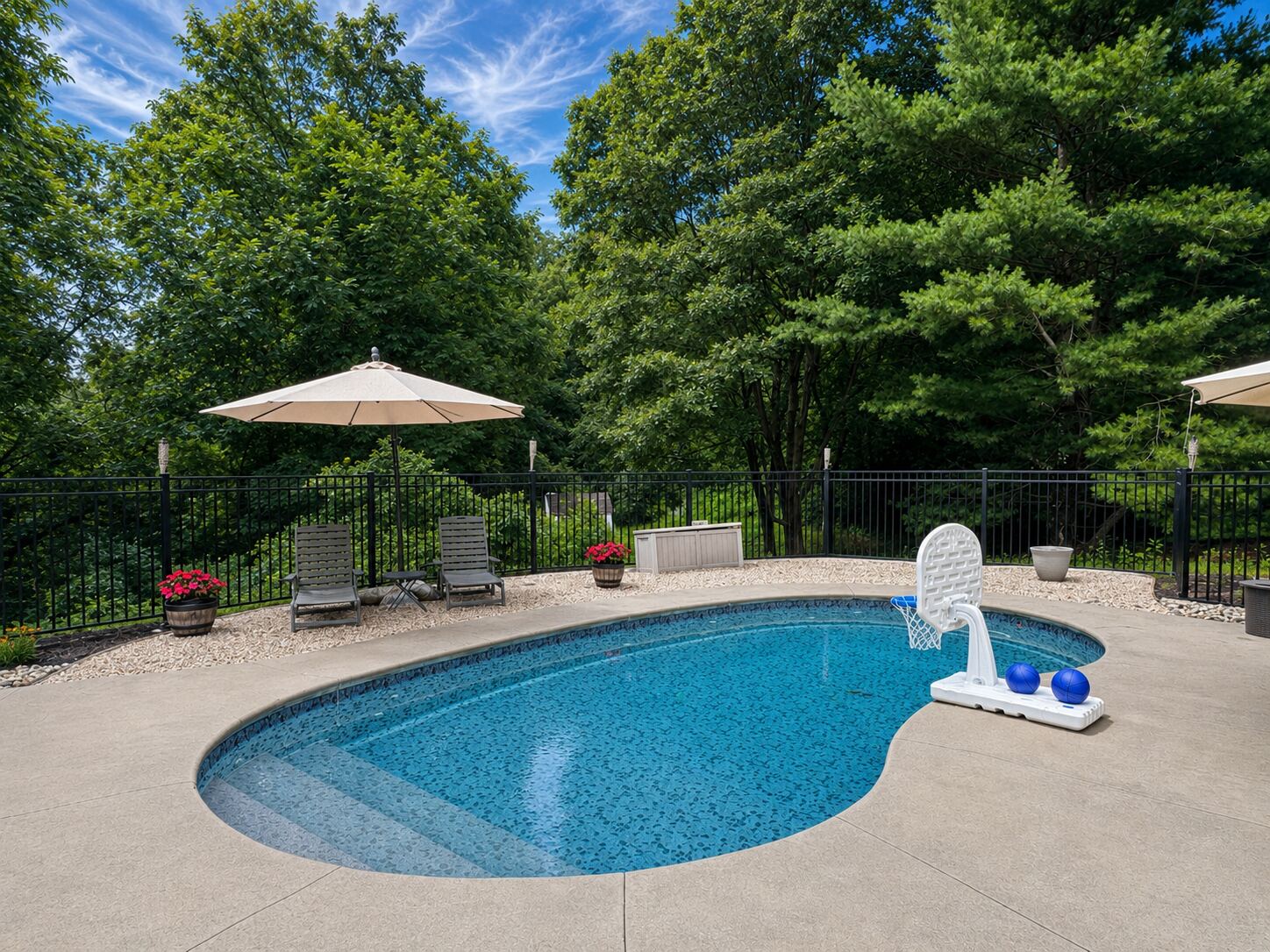 Fenced in-ground pool with concrete surround, umbrellas, lounge chairs, and a poolside basketball hoop